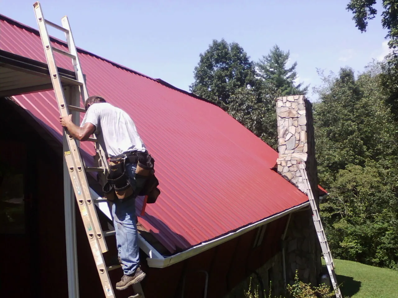 Roofer on a ladder beside a red metal roof and stone chimney — Soffit & Fascia Repair by Roofing Brothers in Storrs Mansfield